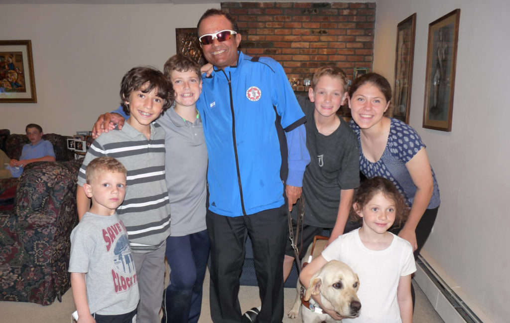 A group of smiling children and a man in sunglasses and a blue jacket stand together indoors. A yellow Labrador guide dog sits in front. There is a couch, brick fireplace, and framed pictures in the background.