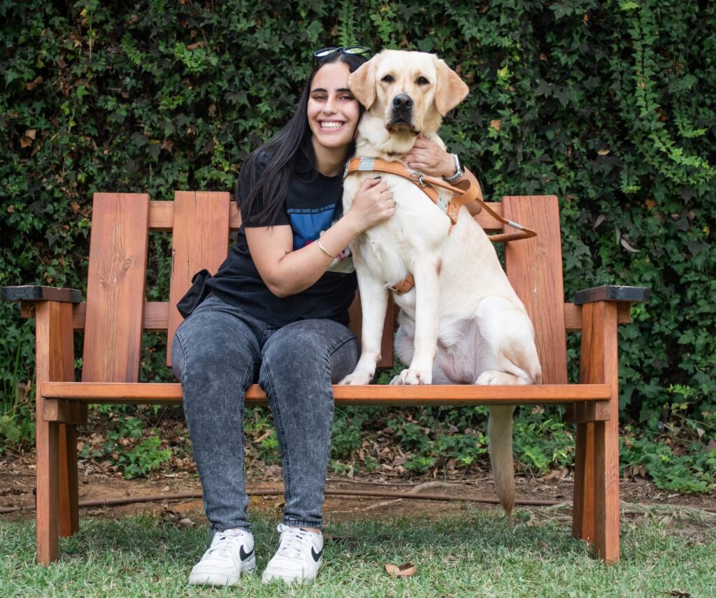 A woman sits on a wooden bench outdoors, smiling and hugging a yellow Labrador Retriever wearing a harness. Green foliage fills the background.
