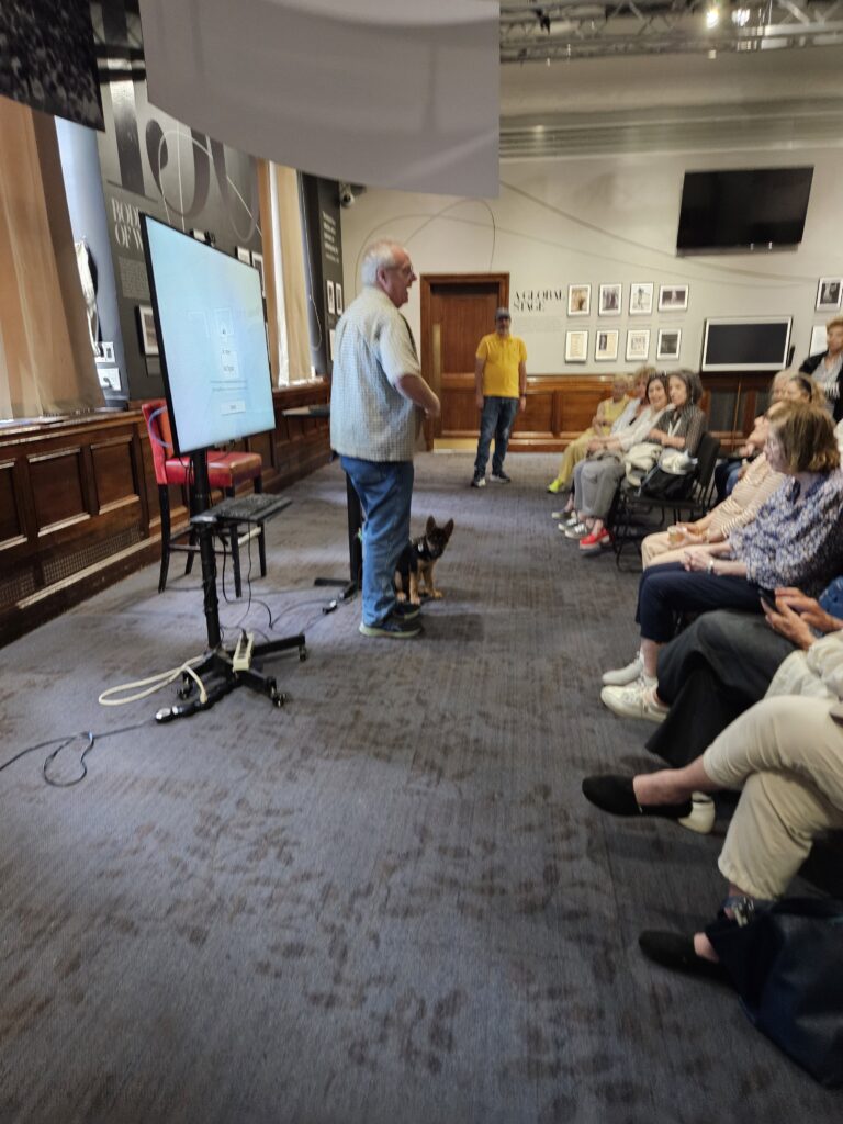 A man stands at the front of a room with a dog, speaking to a seated audience. Theres a large screen beside him, and framed photos hang on the walls in the background.