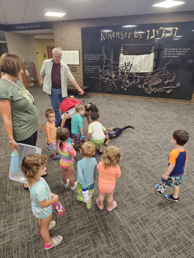 A group of young children and adults watch a dog interacting with kids in front of a Remember Us Holocaust memorial display in a building hallway.