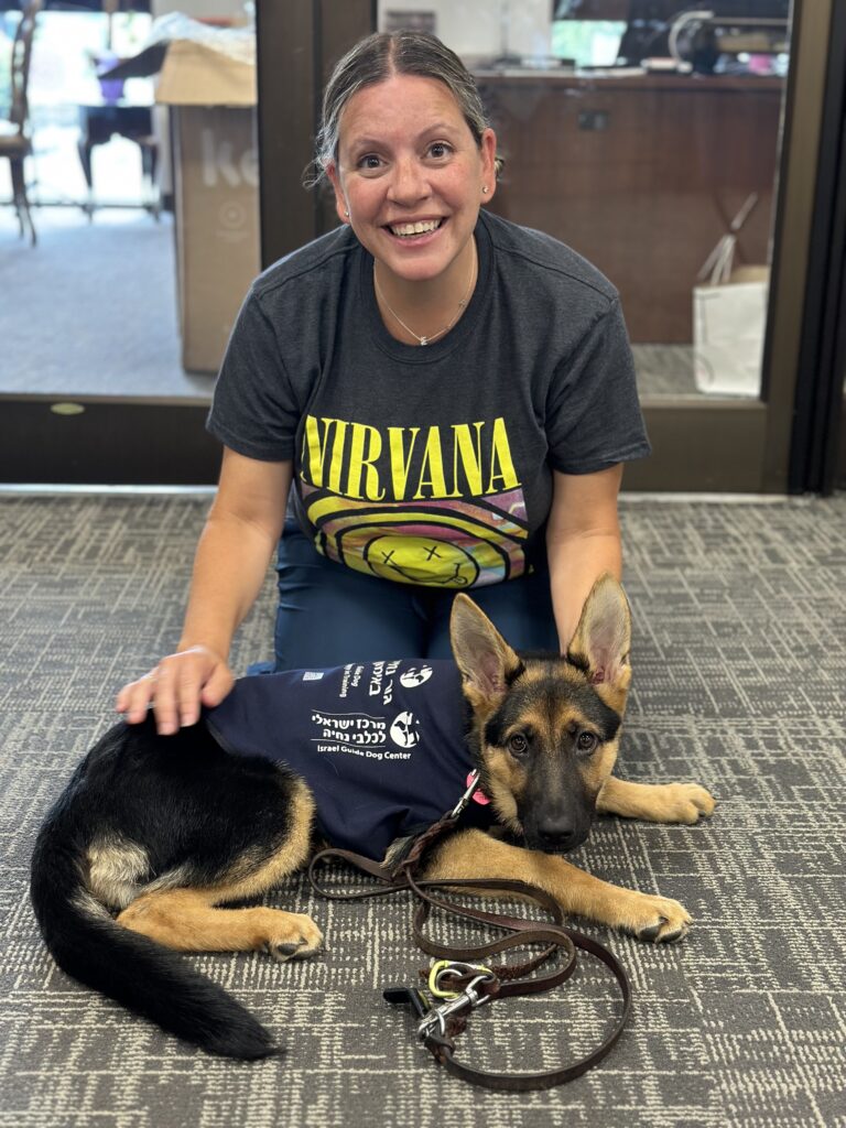 A smiling woman in a Nirvana shirt kneels on the carpet next to a German Shepherd puppy wearing a service dog vest. The puppy is lying down, and a leash is on the floor.