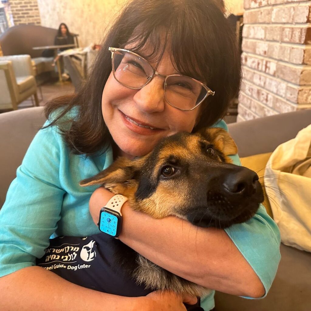 A woman with glasses and a blue shirt hugs a German Shepherd puppy wearing a vest. The woman is smiling, and they are indoors with brick walls and cozy furniture in the background.