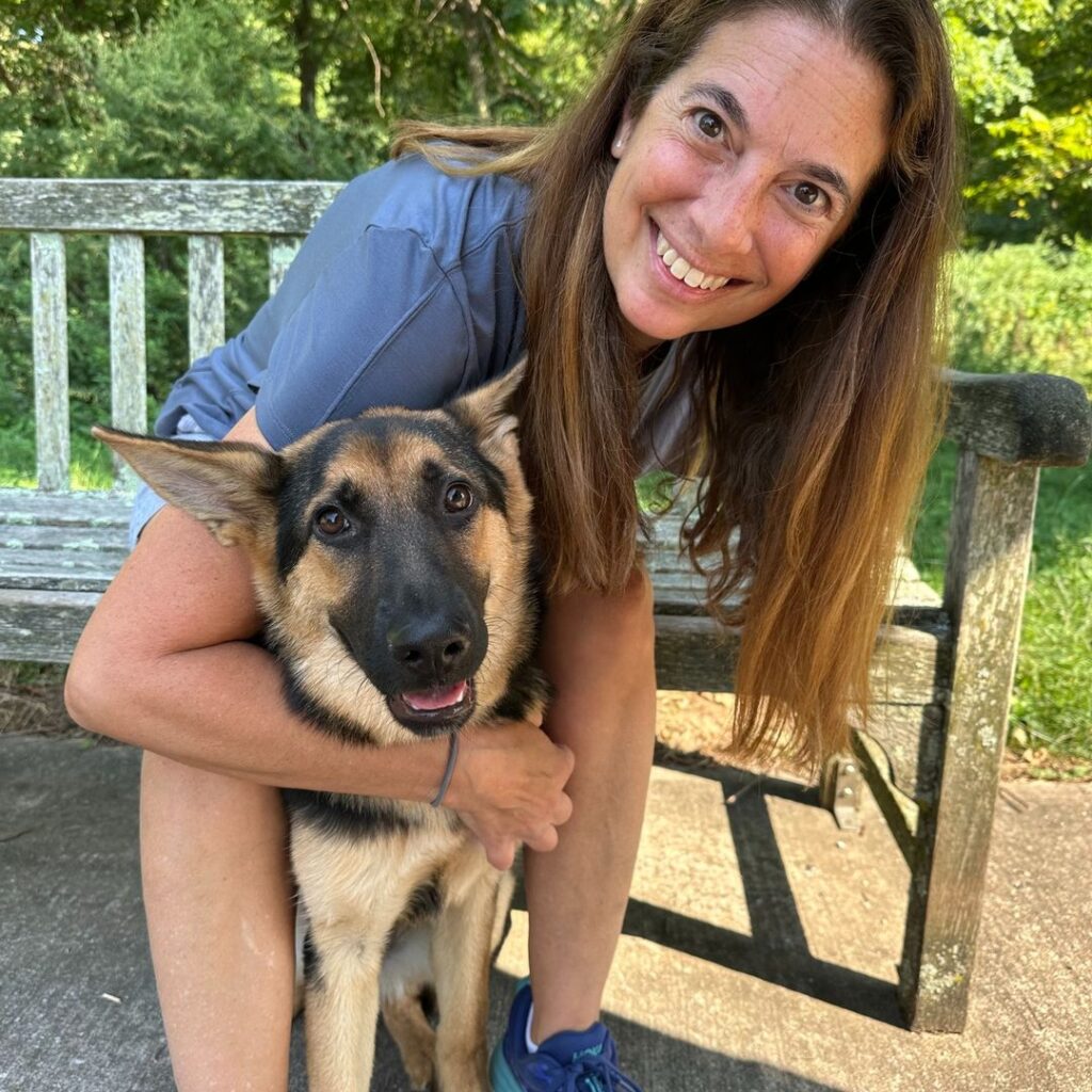 A smiling woman with long brown hair hugs a German Shepherd dog while sitting on a wooden bench outdoors on a sunny day.