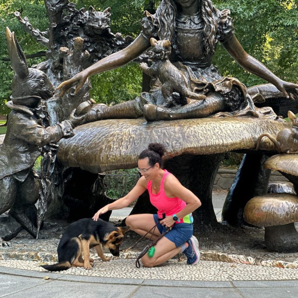 A woman in athletic attire kneels next to a German Shepherd puppy in front of a large Alice in Wonderland-themed bronze statue in a park.
