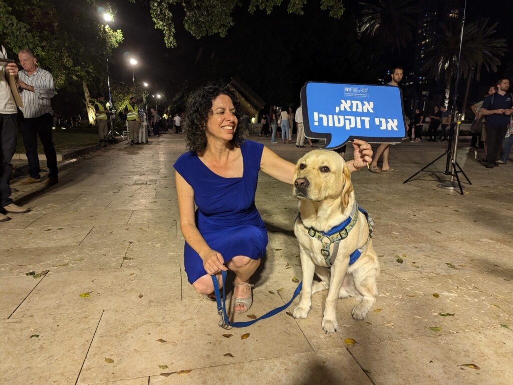 A woman in a blue dress kneels next to a guide dog wearing a harness. She holds a sign in Hebrew above the dog. People stand in the background on a paved area at night.