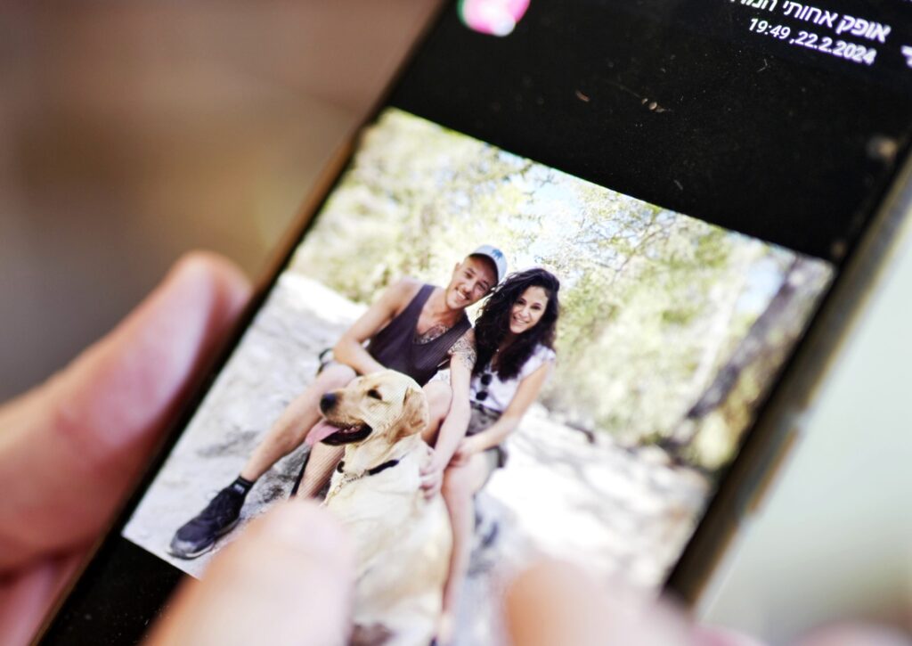 A close-up of a phone screen shows a photo of two people smiling outdoors with a yellow dog, as someone holds and looks at the phone.