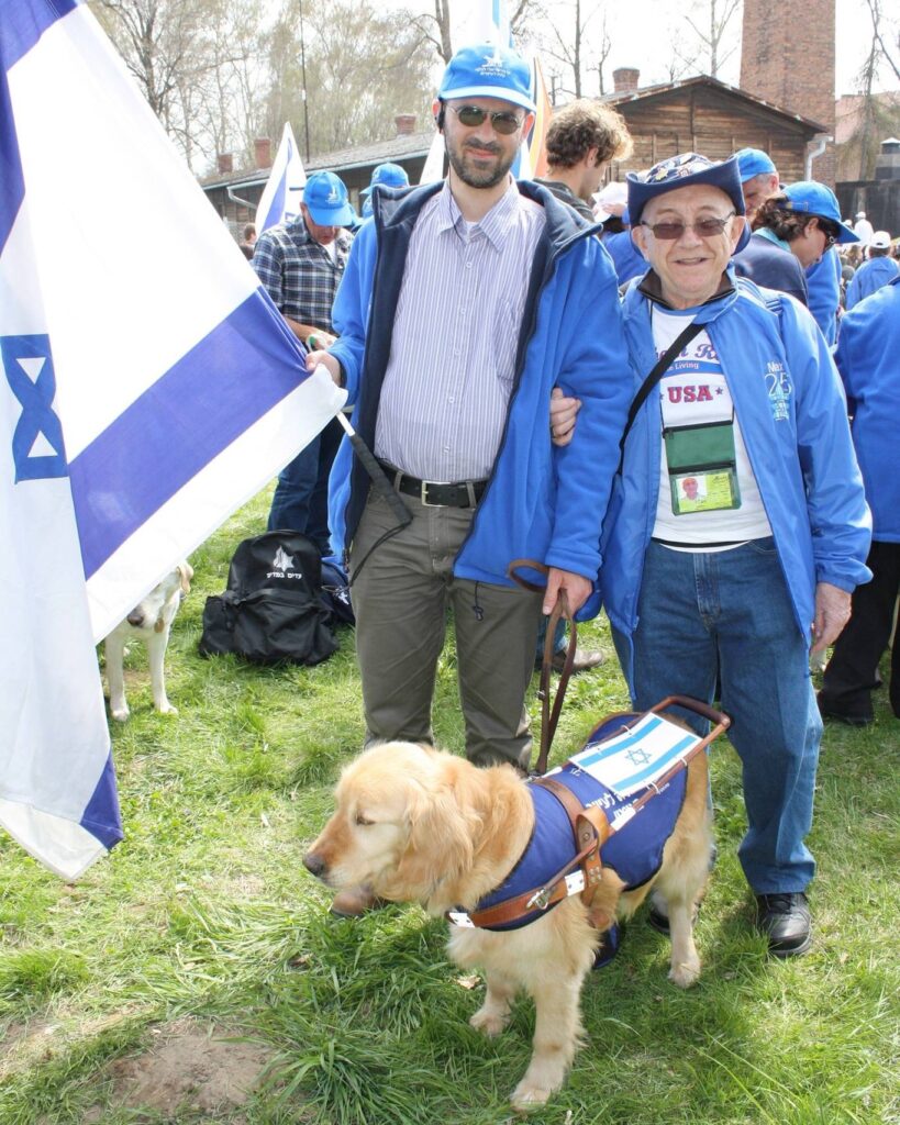 Two men stand on grass; one holds an Israeli flag and the other, wearing a USA badge, holds a guide dogs harness. The dog wears a vest with the Israeli flag. People and buildings are in the background.