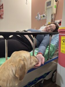 A woman lies in a hospital bed smiling, reaching out to touch a yellow dogs nose. Medical equipment and a yellow biohazard container are visible in the room.