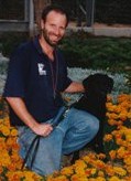 A man kneels in a garden of orange flowers, smiling at the camera while holding a black dog on a leash.