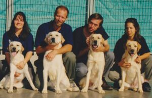 Four people kneel next to four yellow Labrador retrievers, each with a handler. They are outside in front of a corrugated turquoise fence, smiling at the camera.