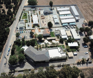 Aerial view of a walled compound with several buildings, parking lots, trees, and landscaped areas, surrounded by fields and a road running along one side.