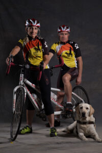Two cyclists in matching jerseys and helmets pose with a tandem bike; a yellow Labrador retriever lies on the floor in front of them. The background is dark and plain.