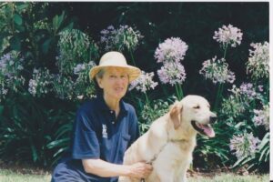 An older woman in a straw hat and navy shirt sits outside on the grass, holding a golden retriever on a leash. Purple flowers and green foliage fill the background.