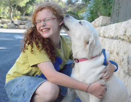 A smiling child with curly red hair and glasses hugs a yellow Labrador retriever, who is licking the childs ear. They are outdoors on a sunny day, next to a stone wall.