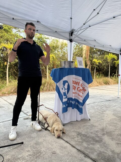 A man gestures while speaking next to a table covered with a blue and white cloth. A guide dog lies on the ground beside him under a white outdoor canopy. Trees and grass are visible in the background.