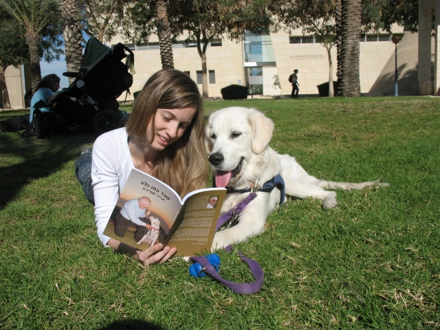 A woman lies on grass next to a golden retriever, reading a book. The dog is looking at the book with its tongue out. Trees, a building, and a stroller are in the background.