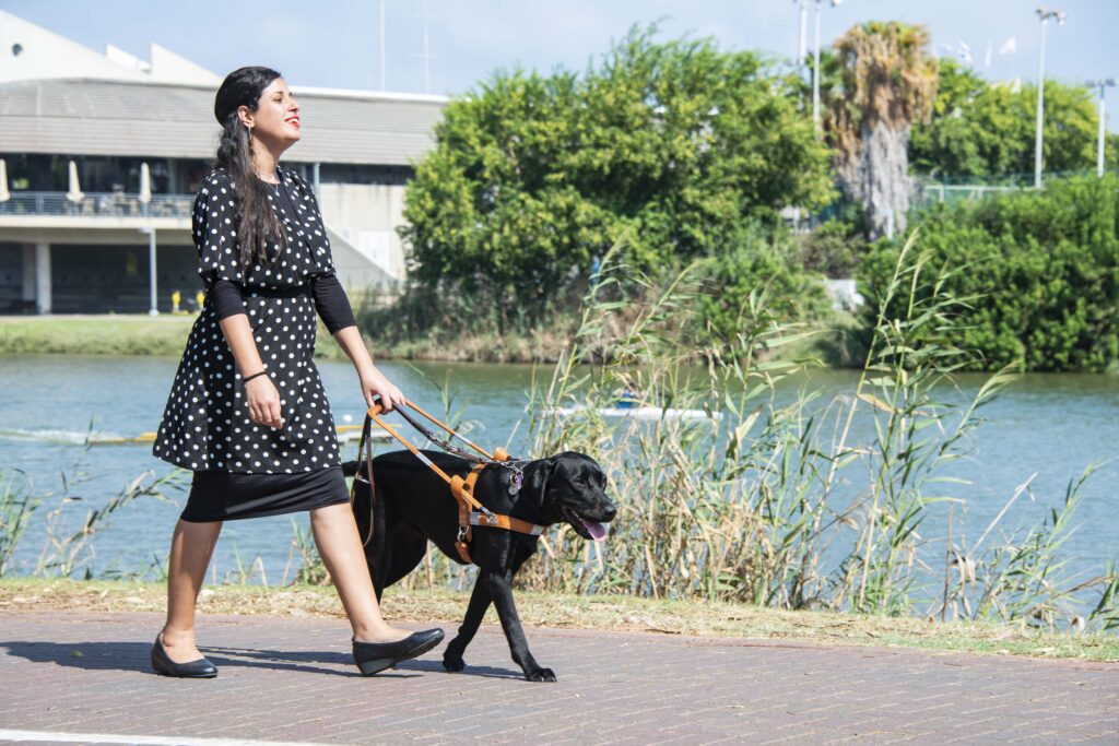 A woman in a black polka-dot dress walks outdoors on a path, guided by a black guide dog on a harness near a river, with trees and a building in the background.