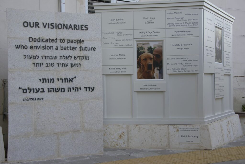 A stone monument features plaques honoring visionaries, with text in English and Hebrew. A central plaque includes a photo of a dog and a person. The dedication reads, Dedicated to people who envision a better future..