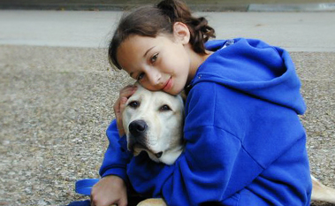 A young girl in a blue hoodie hugs a yellow Labrador retriever while sitting on a gravel surface, both looking content and calm.