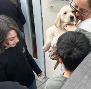 A woman holds a light-colored puppy while two children stand nearby, smiling and interacting with the dog outside a building.