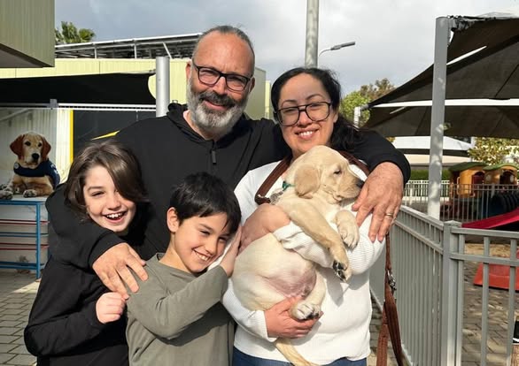 A smiling family of four poses outside, holding a light-colored puppy. Two adults stand behind two children. A dog-themed area and building are visible in the background.