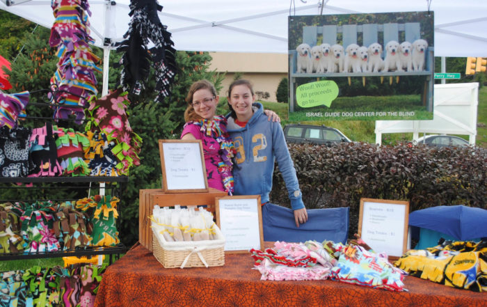 Two smiling people stand behind a colorful vendor table with dog-themed items. A large poster with puppies and signs for a guide dog center are displayed. The scene is outdoors with greenery in the background.